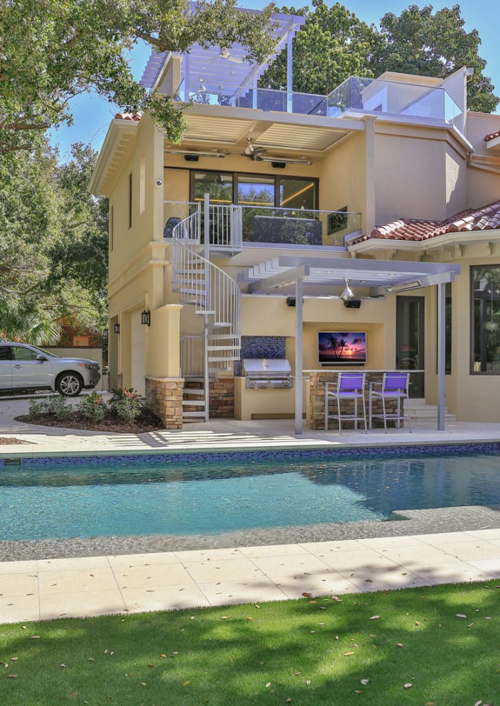 Modern two-story house with a spiral staircase, rooftop deck, pergola-covered patio with grill, outdoor dining area, TV, swimming pool, and green lawn in a sunny backyard setting.