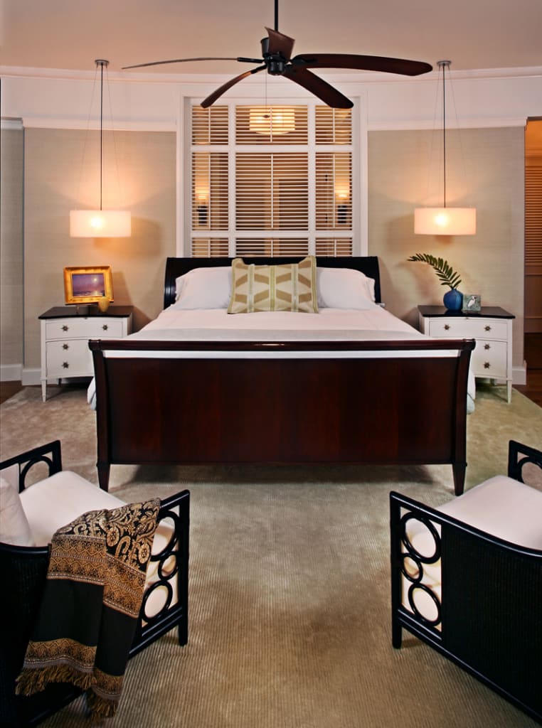 A modern bedroom with a dark wood bed, white bedding, matching nightstands with lamps, armchairs at the foot of the bed, a ceiling fan, and a window with blinds behind the bed.