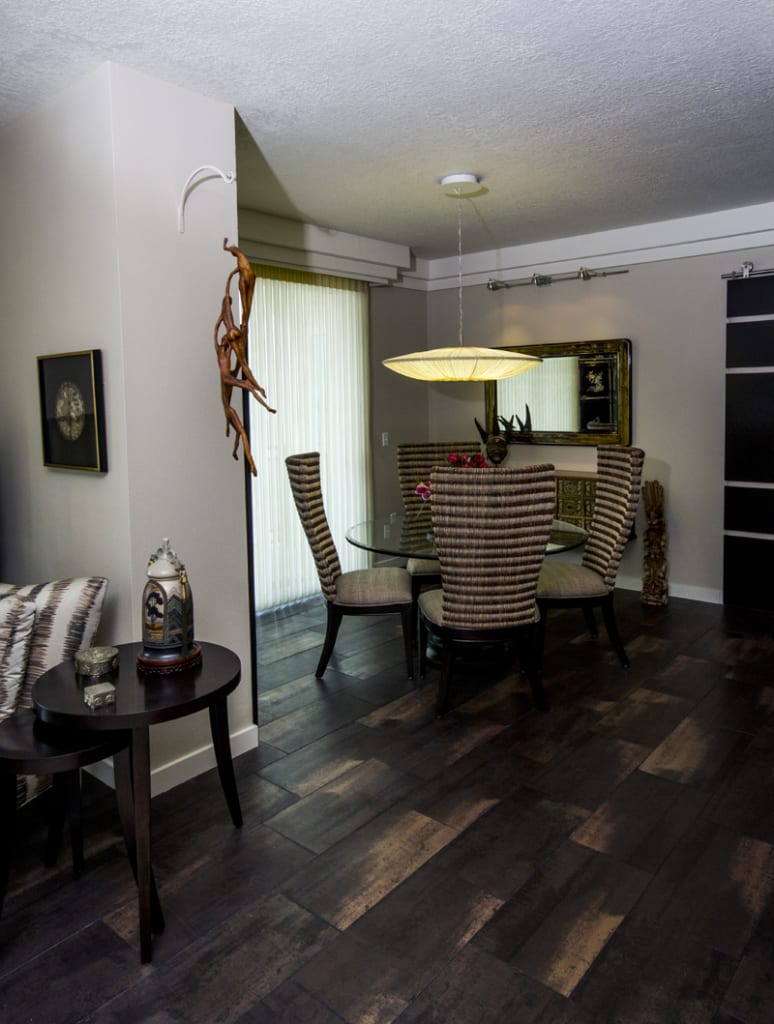 A modern dining room with a round glass table, four striped chairs, a large pendant light above, wall art, and a tall wood sculpture near sheer curtains, with dark wood flooring and neutral walls.