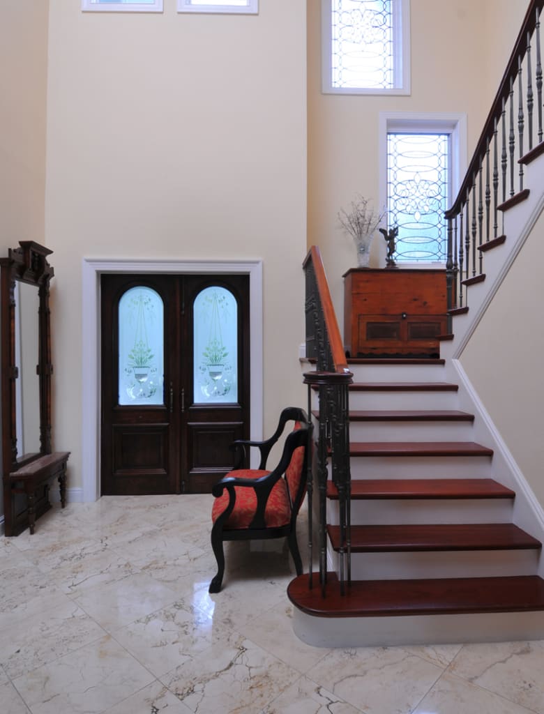 A bright foyer with marble floors, a wooden staircase with metal railings, a large mirror, a cushioned chair, a wooden chest, double doors with frosted glass, and tall windows letting in natural light.