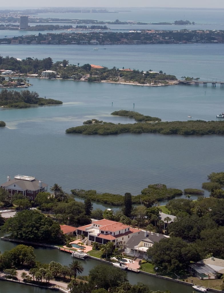 Aerial view of waterfront homes surrounded by lush greenery and water, with small islands and a larger landmass visible in the background under a clear sky.