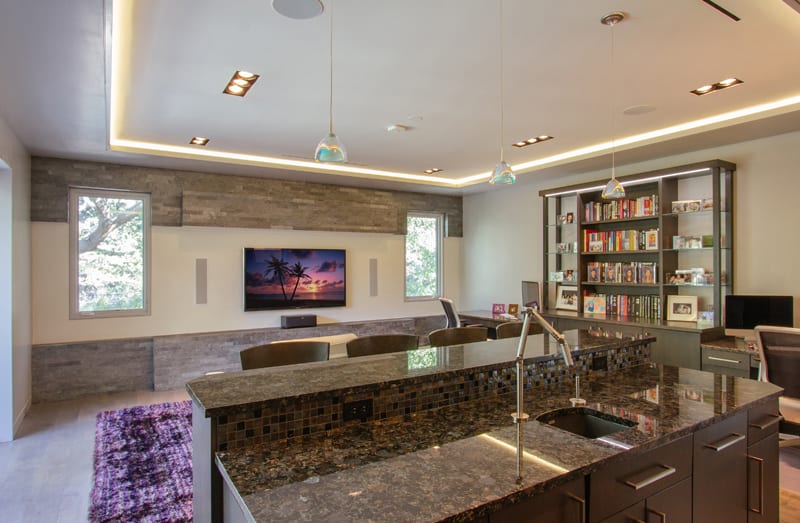 Modern open-concept living space with a kitchen island in the foreground, a TV mounted on the wall, bookshelves, a desk, and large windows letting in natural light.