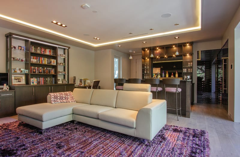 Modern living room with a white sectional sofa on a purple rug, a bookshelf and cabinet on the left, and a home bar with high chairs and pendant lights in the background. The space has sleek decor and warm lighting.
