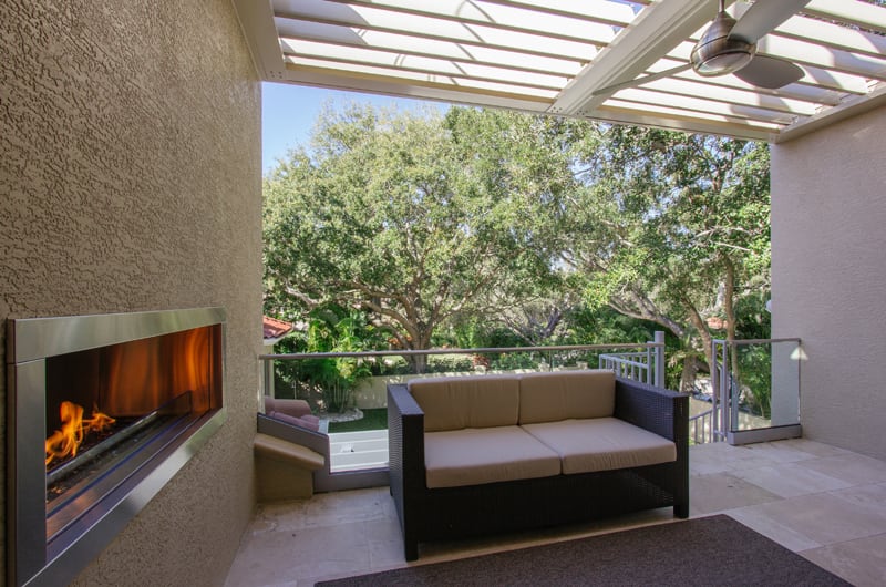 A modern patio with a built-in wall fireplace, a wicker sofa with light cushions, and a glass railing overlooking a green yard with large trees. The patio has a slatted roof letting in sunlight.