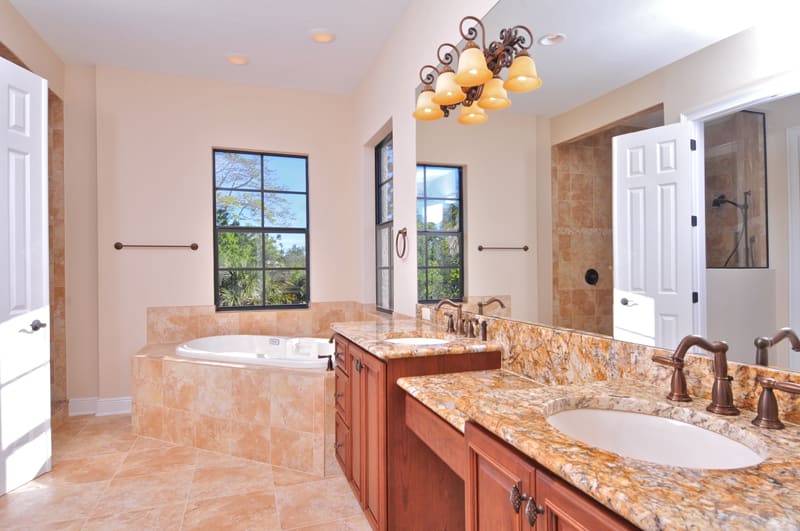 A spacious bathroom with a large soaking tub by two windows, double sinks with marble countertops, wooden cabinets, and a walk-in shower with glass door. The room has beige tiles and warm lighting fixtures.