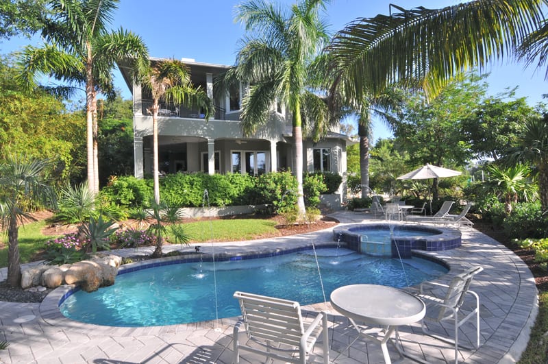 A tropical backyard features a small pool with fountains, surrounded by palm trees and lush greenery. There are lounge chairs and a table on the pool deck, with a two-story house in the background.