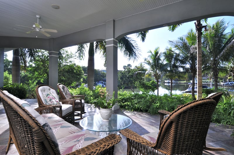 A covered patio with wicker furniture and a glass table overlooks a lush garden with palm trees and a view of a marina with boats on the water in the background.