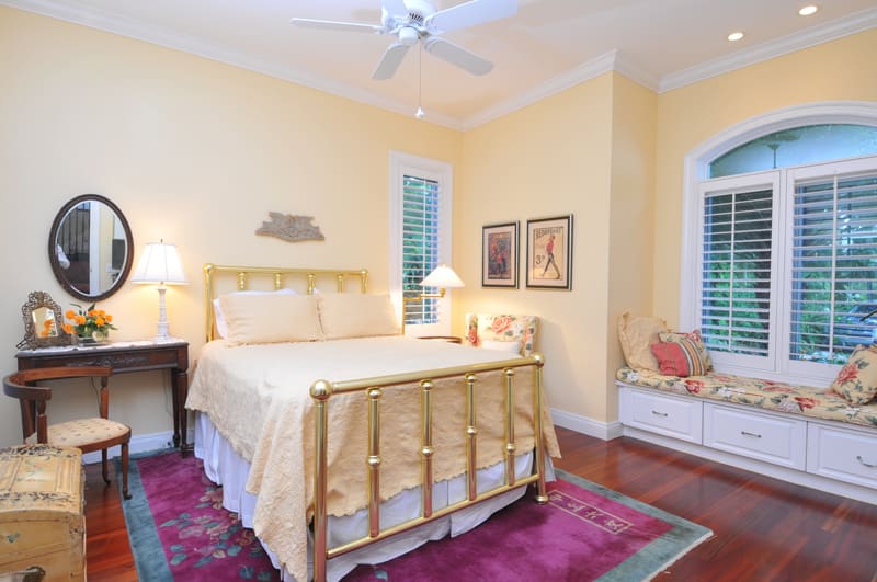 A cozy bedroom with a brass bed, yellow bedding, and matching walls. There is a vintage vanity with a round mirror, a window seat with cushions, and framed art on the walls. Hardwood floors and a ceiling fan complete the room.