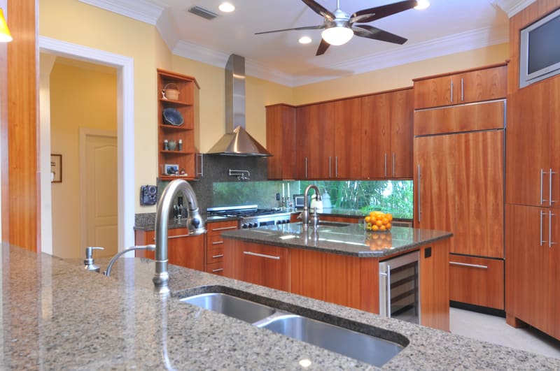 Modern kitchen with wooden cabinets, stainless steel appliances, large island with a sink, granite countertops, a window above the stove, and a bowl of oranges on the island. Ceiling fan and recessed lighting overhead.