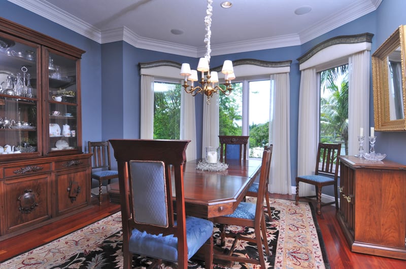 A traditional dining room with a wooden table, six blue-cushioned chairs, a chandelier, a patterned rug, and a china cabinet. Large windows with white curtains show green trees outside.
