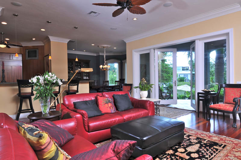 A spacious living room with red leather sofas, a black ottoman, and colorful pillows. Large windows and glass doors open to a patio with greenery. The room includes a dining area, kitchen, and elegant light fixtures.
