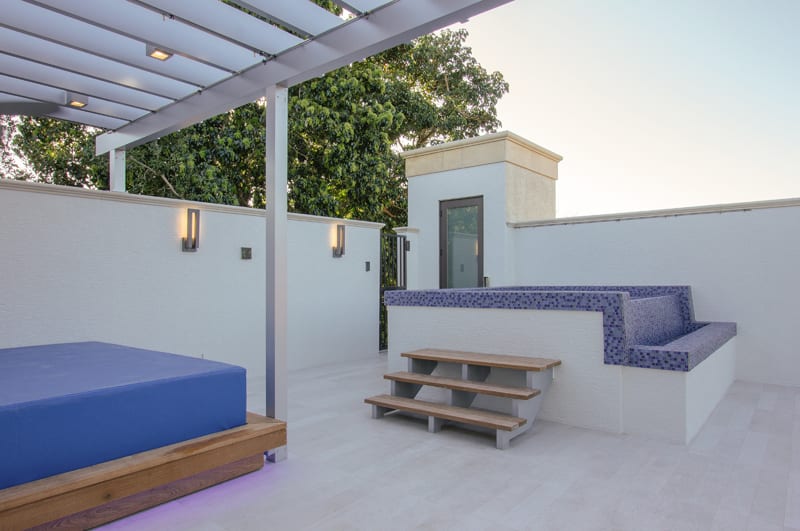 Modern rooftop patio with white walls, a pergola, built-in seating with blue cushions and mosaic tile, wooden steps, and outdoor wall lights; trees and blue sky in the background.
