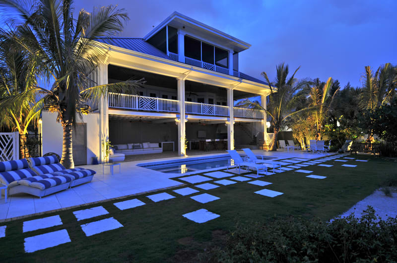 A modern two-story house with large balconies and outdoor seating is illuminated at dusk. A swimming pool is surrounded by lounge chairs, palm trees, and stone pavers set in the grassy yard.