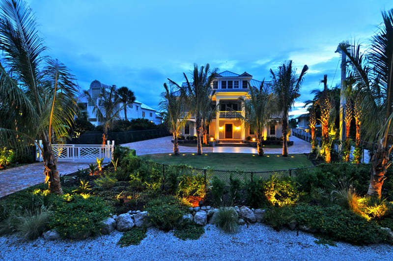 A large, elegant house with three stories is illuminated at dusk, surrounded by palm trees, manicured landscaping, and a circular driveway, creating a tropical, inviting atmosphere.