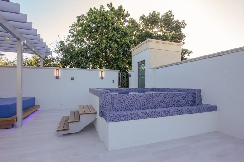 Modern outdoor patio with a white tile floor, a built-in bench covered in purple mosaic tiles, wooden steps, white walls, a pergola, and trees in the background.