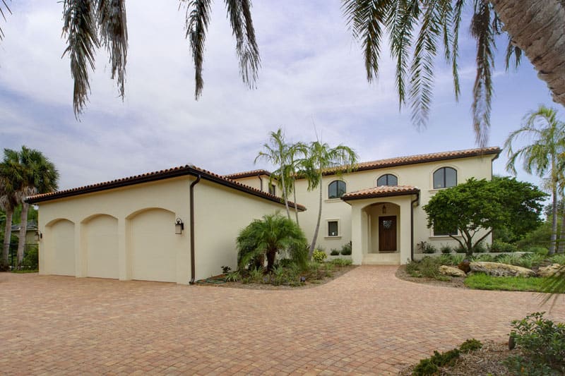 A two-story Mediterranean-style house with a red-tiled roof, light stucco walls, three-car garage, arched windows, and a paved driveway, surrounded by palm trees and greenery under a partly cloudy sky.