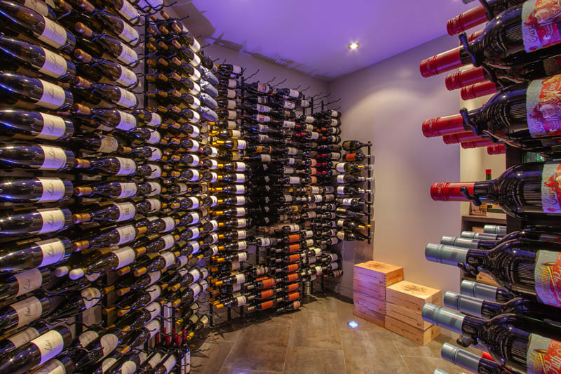 A wine cellar with walls lined with racks holding numerous bottles of wine, some with red and some with white labels. Wooden wine crates are stacked on the floor beneath soft overhead lighting.
