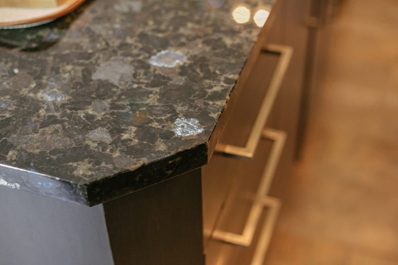 Close-up of a dark, polished stone countertop with a beveled edge above a set of wooden drawers featuring modern metallic handles. Warm, soft lighting reflects off the surface.