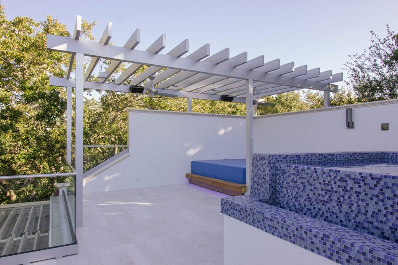 A modern outdoor terrace with a white pergola, a blue-cushioned daybed, and a tiled hot tub featuring blue mosaic tiles, surrounded by white walls and trees in the background.