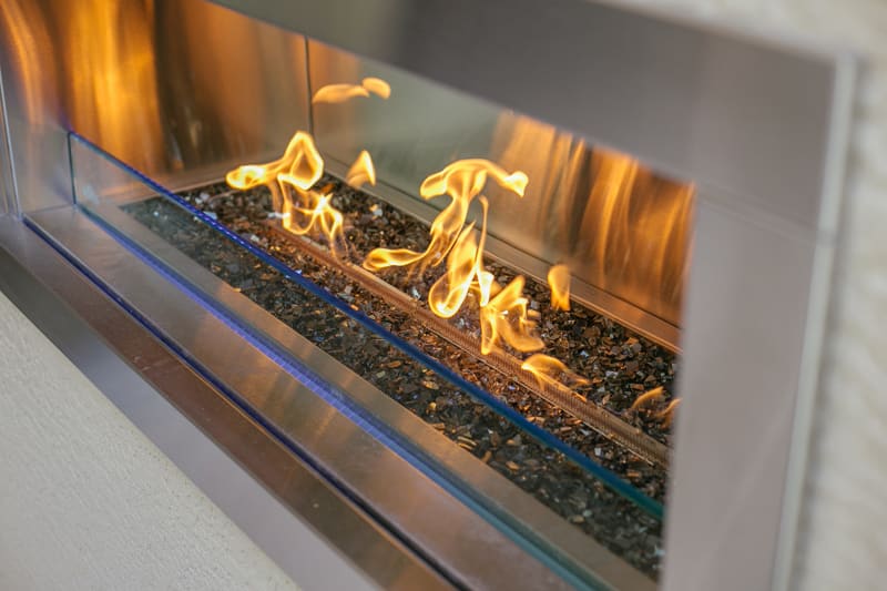 Close-up of a modern, rectangular gas fireplace with clean metal edges and decorative glass stones, showing bright yellow-orange flames inside.