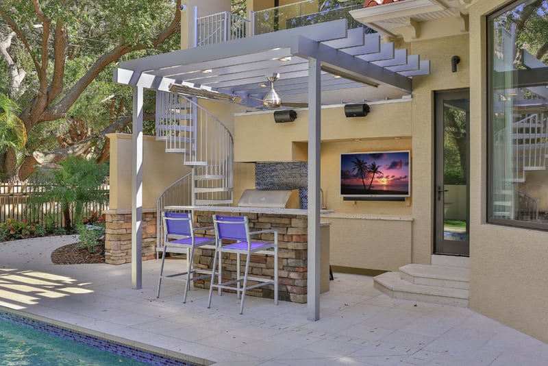 Outdoor patio with a pergola, spiral staircase, bar stools, built-in grill, and a wall-mounted TV showing a beach scene next to a pool, surrounded by trees and greenery.