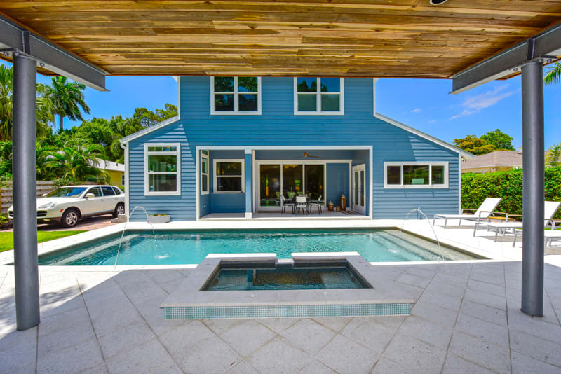 View of a modern blue house with large windows, a covered patio, and a backyard swimming pool with a hot tub. There are lounge chairs and a car parked on the left, with greenery in the background.