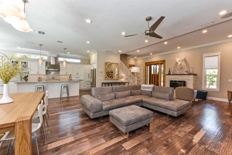 Spacious open-concept living area with a gray sectional sofa, modern kitchen, wooden dining table, and hardwood floors. A staircase, fireplace, and mounted animal head decorate the room. Ceiling fans and recessed lighting provide illumination.