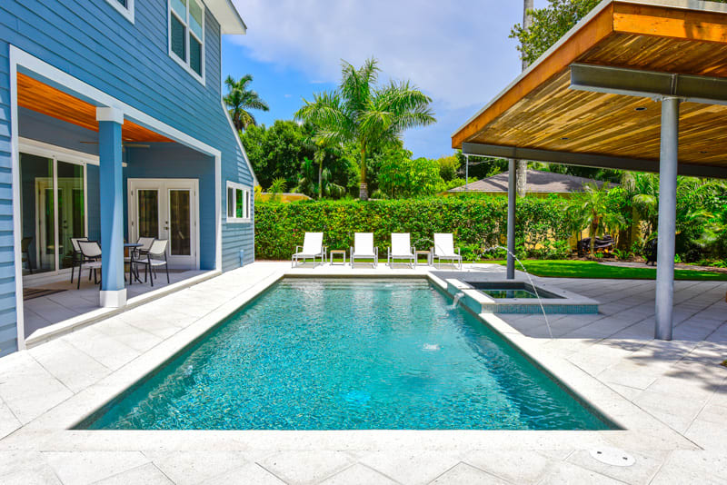 A modern backyard with a rectangular swimming pool, a hot tub, several white lounge chairs, a covered patio area, and tropical landscaping with palm trees and greenery.