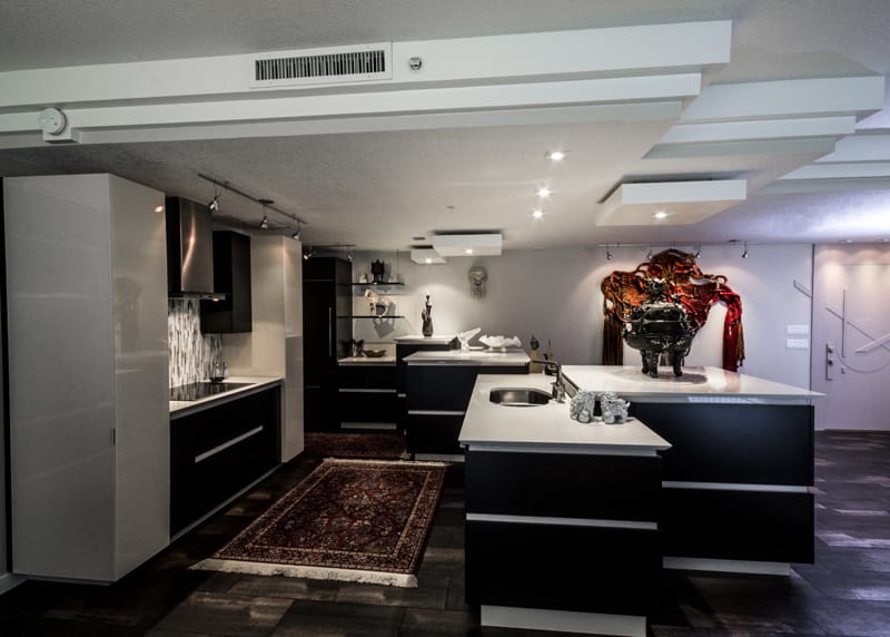 Modern kitchen with black and white cabinetry, a red patterned rug, and contemporary decor, including sculptures and wall art. The space features sleek counters, a built-in oven, and recessed lighting.