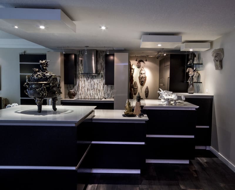 Modern kitchen with dark cabinets, white countertops, and contemporary sculptures on counters. Wall displays include tribal masks and artistic sculptures, creating a gallery-like atmosphere with dramatic lighting.