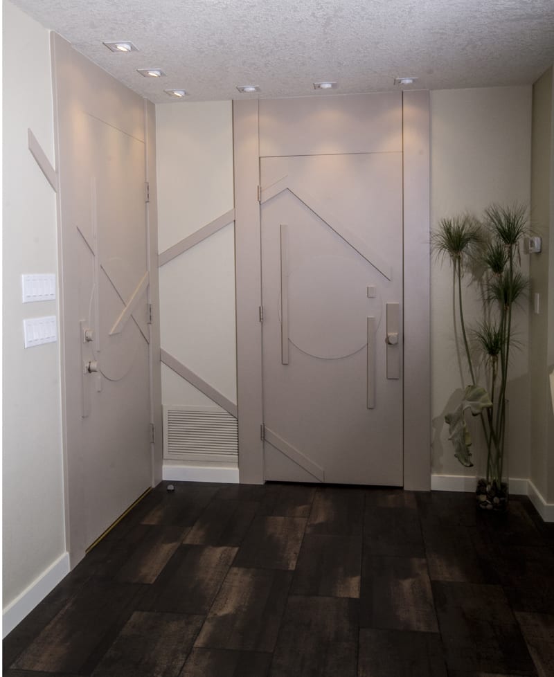 Modern entryway with two geometric-patterned beige doors, dark wood flooring, recessed ceiling lights, light-colored walls, and a tall green plant in the corner by a thermostat and three light switches.