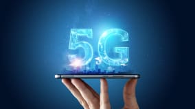 Eine Hand hält ein Smartphone, über dem in leuchtendem, transparentem Blau die Zahlen und Buchstabe "5G" schweben. Die Szene ist vor einem dunklen Hintergrund dargestellt, wobei ein sanfter Lichtschein von den Zahlen ausgeht und die Umgebung beleuchtet. Im Hintergrund ist eine abstrakte Silhouette einer Stadt zu erkennen.