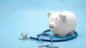 Ein weißes Sparschwein steht auf einem blauen Hintergrund. Daneben liegt ein blaues Stethoskop, das den Eindruck vermittelt, Finanzthemen im Gesundheitsbereich anzusprechen.