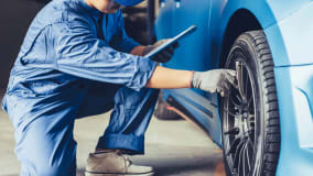 Ein Mechaniker in blauer Arbeitskleidung kniet neben einem blauen Auto und inspiziert ein Rad. Er hält ein Tablet in der rechten Hand und benutzt die linke Hand, um die Radfelge zu berühren. Im Hintergrund sieht man Teile der Fahrzeugkarosserie und den Boden einer Werkstatt. Der Mechaniker trägt graue Handschuhe und weiße Sneakers.