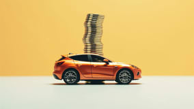 Ein orangenes Modellauto steht auf einer ebenen Oberfläche. Auf dem Dach des Autos ist ein großer Stapel goldener Münzen platziert. Der Hintergrund ist einfarbig in einem hellen Gelbton gehalten.