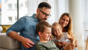 Eine Familie sitzt zusammen auf einem beigen Sofa in einem hellen Wohnzimmer. Der Vater trägt ein jeansblaues Hemd und legt seinen Arm um seine Kinder. Ein Junge im Vordergrund hält ein Tablet in den Händen, während ein kleines Mädchen neben ihm sitzt. Im Hintergrund ist die Mutter zu sehen. Licht fällt durch große Fenster in den Raum.