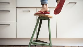 Eine Person mit roten Flip-Flops und hochgekrempelten Jeans steht auf der Sitzfläche eines grünen, vintage Metallhockers in einer Küche mit weißen Schränken und Holzboden. Der Hocker wirkt wackelig und es scheint, als würde die Person gerade den Hocker verlassen oder daraufsteigen.