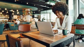 Eine Frau mit lockigem Afro-Haar sitzt in einem modernen Café mit gemütlicher Einrichtung auf einem hölzernen Stuhl vor einem Laptop an einem Holztisch. Neben dem Laptop steht eine Tasse. Im Hintergrund sind weitere Gäste und stilvolle, warme Beleuchtung sichtbar.