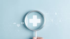 Eine Hand hält eine Lupe, durch die ein weißes Kreuz auf einem blauen Hintergrund zu sehen ist. Um das Kreuz herum befinden sich transparente medizinische Symbole und hexagonale Muster, die ein digitales Gesundheitskonzept darstellen.