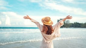 Frau mit langen, braunen Haaren steht am Strand, trägt einen Strohhut und eine weiße Bluse. Sie breitet ihre Arme weit aus, als würde sie die frische Meeresbrise und die Freiheit des offenen Himmels genießen. Im Hintergrund sind das Meer und ein bewaldetes Ufer zu sehen. Der Himmel ist blau mit einigen weißen Wolken.