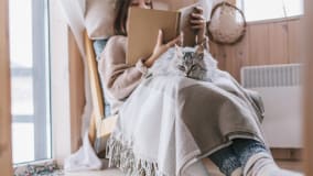 Eine Frau sitzt gemütlich auf einem Sessel in einem Holzraum und liest ein Buch. Sie ist mit einer grauen Decke zugedeckt, auf der eine pelzige graue Katze liegt. Die Frau trägt warme Wintersocken. Neben ihr ist ein Fenster, durch das Tageslicht in den Raum fällt.