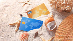 Zwei Kreditkarten, eine in Blau und eine in Gold, liegen im Sand, umgeben von verschiedenen Muscheln und Seesternen. Im Hintergrund ist ein Stück Koralle zu sehen, und an der Seite ragt ein Sonnenschirm aus Bast ins Bild.