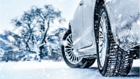 Nahaufnahme eines Autos, das auf einer schneebedeckten Straße geparkt ist. Die Reifen sind klar erkennbar und mit Schnee bedeckt. Im Hintergrund sind Schneeflocken sichtbar, die fallen, und ein schneebedeckter Baum vor einer unscharfen Winterlandschaft.