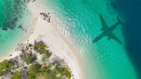 Ein Luftbild einer tropischen Küste mit weißem Sand, klarem türkisfarbenem Wasser und grüner Vegetation auf der Insel. Im Meer ist der deutliche Schatten eines Flugzeugs zu erkennen, das über die Landschaft fliegt. Ein paar verstreute Bauten und Sonnenschirme sind auf dem Strand erkennbar.