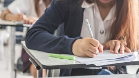 Eine junge Frau sitzt an einem Schreibtisch in einem Klassenzimmer und schreibt mit einem Bleistift auf ein Blatt Papier. Vor ihr liegt ein grüner Stift auf dem Tisch. Im Hintergrund sind verschwommen weitere Personen zu sehen, die ebenfalls an Tischen sitzen und schreiben.