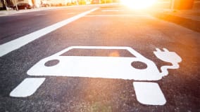 Weiße Straßenmarkierung in Form eines Autos mit einem Stromstecker-Symbol auf einem Asphaltparkplatz, bei tiefstehender Sonne und warmem Licht im Hintergrund.
