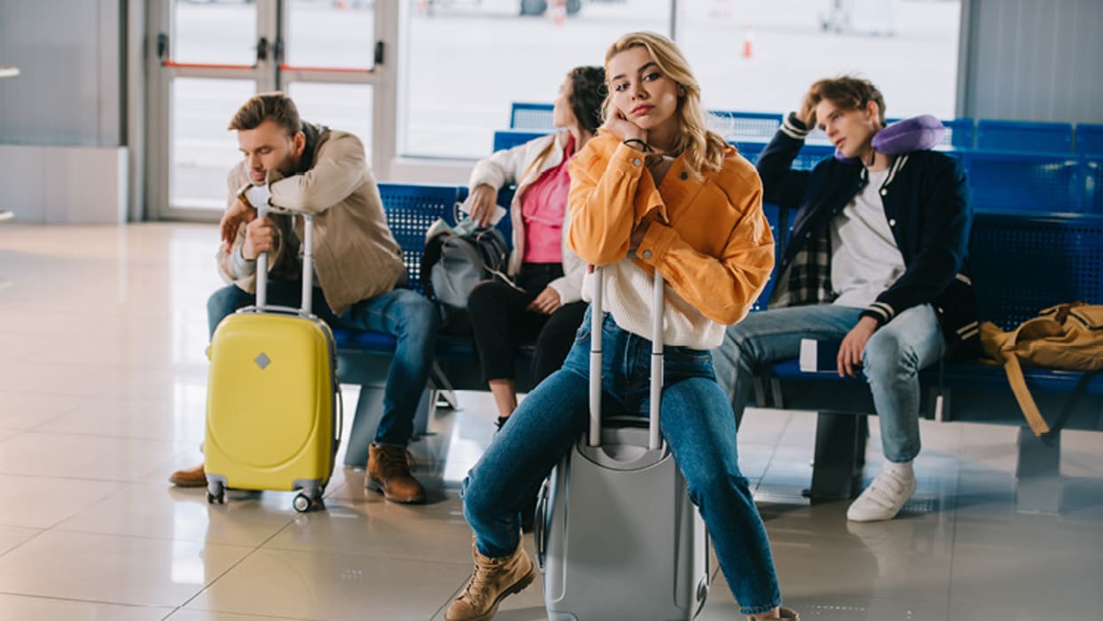 ALT-Tag: Vier Personen sitzen in einem Wartebereich eines Flughafens. Sie wirken gelangweilt und müde. Eine Frau in einer orangefarbenen Jacke sitzt auf einem grauen Rollkoffer, während eine andere Person einen gelben Koffer vor sich hält. Im Hintergrund sind blaue Sitzbänke und Glastüren zu sehen, die den Blick auf das Flughafenvorfeld freigeben.