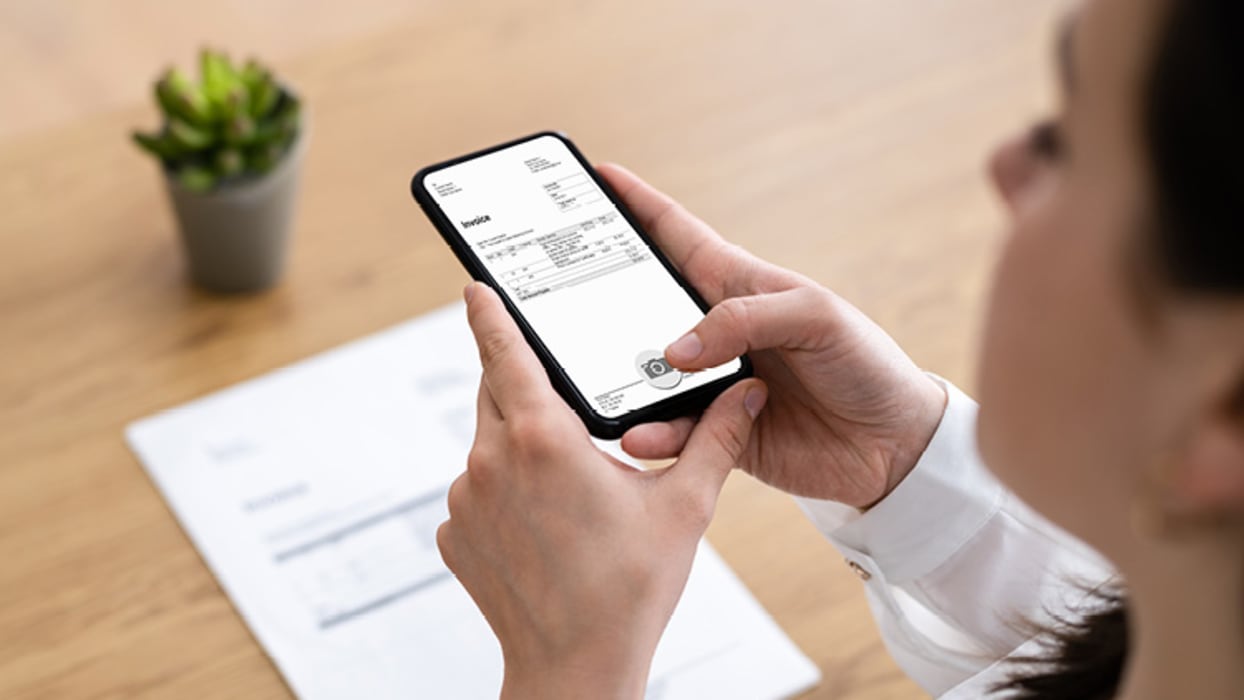 ALT-Tag: Eine Person fotografiert mit einem Smartphone ein Dokument oder eine Rechnung. Das Dokument liegt auf einem Holztisch, daneben ist eine kleine grüne Pflanze in einem Topf zu sehen. Die Person trägt ein weißes Hemd, und ihr Gesicht ist unscharf gemacht.