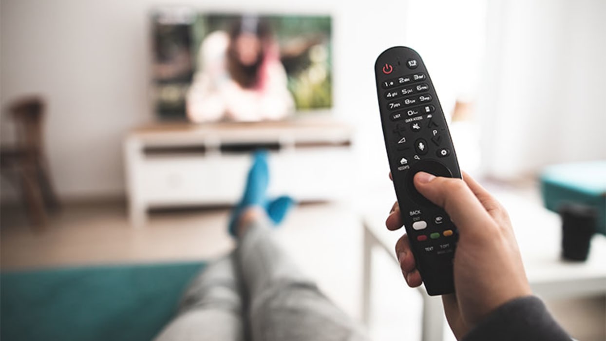 Eine Person hält eine schwarze Fernbedienung in der Hand, während sie auf einem Sofa sitzt. Im Hintergrund ist ein unscharfer Fernseher zu sehen, auf dem eine Person dargestellt ist. Der Raum ist hell und modern eingerichtet, mit einem niedrigen weißen Fernsehschrank.