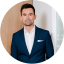ALT-Tag: Eine Person in einem eleganten, dunkelblauen Anzug und weißem Hemd steht vor einem Hintergrund, der teilweise aus einer hölzernen Wand und einer weißen Glasfläche besteht. Das Gesicht der Person ist unscharf gemacht.